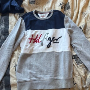 Grå och marinblå sweatshirt från Tommy Hilfiger Denim - Säljer en snygg sweatshirt från Tommy Hilfiger Denim i grått, vitt och marinblått. Tröjan har broderad logga framtill och rund halsringning med ribbade muddar. Perfekt för en avslappnad stil.