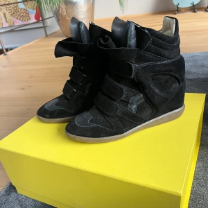 Svarta sneakers med kilklack från Isabel Marant - Säljer ett par svarta sneakers med kilklack från Isabel Marant. Skorna har en cool design med både mocka och skinn, samt breda kardborreband och rund tå. Perfekta för dig som vill ha både stil och höjd i vardagen.