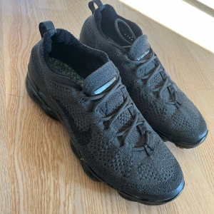 Nike air Vapormax  - Svarta Nike Vapormax Flyknit sneakers med stickad ovandel och platt sula. Skorna har snörning och en modern, sportig look med synlig dämpning i sulan. Perfekta för dig som gillar stilrena och bekväma sneakers.