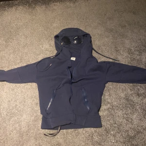 Marinblå hoodie med goggles från C.P. Company - Tja tänkte nu sälja min cp company kofta eftersom den blivit för liten. Inte använd så mycket,men tecken finns att den är använd som är på en av fickorna, men inte så märkvärdigt. Storleken är S men är liten för storleken passar mer till xs skulle jag säga, men det är än väldigt fin kofta skulle säga skicket är 8-10 på den!!
