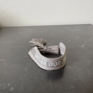 Grått armband från Dior - Säljer ett snyggt grått armband från Dior med broderad logga och tofsar i ändarna. Perfekt accessoar för att lyfta din outfit med en lyxig touch.