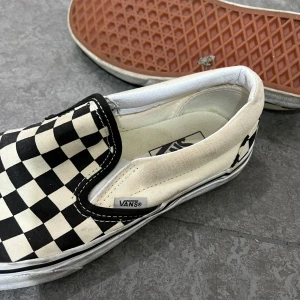 Vans slip-on checkerboard - Klassiska Vans slip-on sneakers med svartvitt rutigt mönster. Skorna har vit sula och elastiska paneler på sidorna för enkel påtagning. Perfekta för en avslappnad och cool stil.
