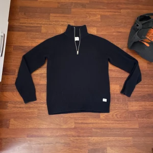 Mörkblå stickad tröja från Jack & Jones - Säljer en stilren Mörkblå stickad tröja från Jack & Jones med dragkedja vid halsen. Perfekt för kyligare dagar och passar till det mesta. Tröjan har långa ärmar och en diskret logga vid nederkanten.