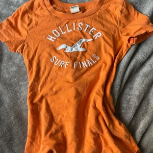 Orange t-shirt från Hollister - Orange t-shirt från Hollister med vit text och logga på bröstet. Klassisk rund halsringning och korta ärmar. Perfekt för sommaren! Bra om du gillar y2k stilen! Den är i helt okej skick inga fläckar eller hål. Är en storlek S men materialet är ganska stretchigt!