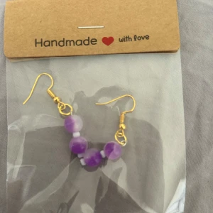 Handgjorda örhängen med lila pärlor - Säljer ett par handgjorda örhängen med två lila pärlor och små vita detaljer. Krokarna är i guldfärg och ger en fin kontrast till de lila pärlorna. Perfekta för att ge din stil en färgklick!🫶🏼💜