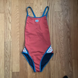 Röd baddräkt från Arena med blå detaljer - Sportig röd baddräkt från Arena med blå kantband och smala axelband. Baddräkten har öppen rygg och inslag av vitt och blått mönster vid höfterna. Perfekt för simträning eller bad.