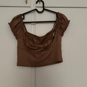 Brun croppad topp med volang och knyt - Säljer en brun croppad topp med volangdetaljer vid ärmarna och en snygg knytning i ryggen. Toppen har korta puffärmar och en söt, romantisk stil. Perfekt för sommardagar eller när du vill känna dig extra fin.