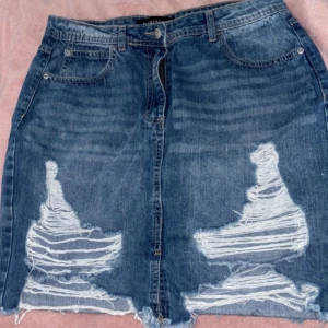 Blå jeanskjol med slitningar från Parisian - Snygg blå jeanskjol från Parisian med rå kant och stora slitningar framtill. Klassisk femficksmodell och knappstängning. 