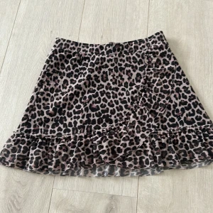 Leopardmönstrad volangkjol från lindex - Superfin kort kjol från Lindex med leopardmönster i svart och beige. Kjolen har volang nertill och omlott-look framtill som ger en trendig vibe. Perfekt för dig som gillar att sticka ut med coola mönster och detaljer.
