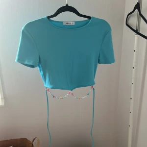 Turkos croppad topp  - Turkos ribbad croppad topp med kort ärm och rund hals. Nedtill finns en unik detalj med snörning och färgglada pärlor. 