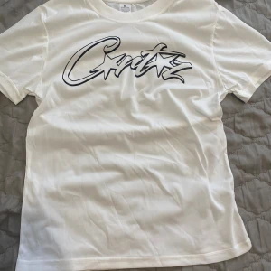 Vit t-shirt med grafiskt tryck Corteiz - Vit t-shirt från Corteiz med ett stort svart grafiskt tryck på bröstet. Klassisk passform med rund hals och korta ärmar. Tillverkad i mjuk bomull som känns skön mot huden. Perfekt för dig som gillar streetwear vibes och enkel stil. Priset kan diskuteras!