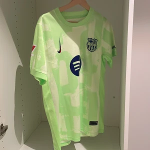 FC Barcelona Raphinha 11 Nike tröja - FC Barcelona matchtröja med Raphinha 11-tryck. Ljusgrön och vit med abstrakt mönster, klubbmärke och Nike-logga på bröstet samt Spotify och UNHCR-tryck. Kortärmad och tillverkad i Dri-FIT-material som andas. Perfekt för dig som älskar Barca och vill sticka ut.