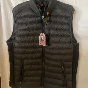 Svart dunväst från Parajumpers - Snygg svart dunväst från Parajumpers med quiltad design och hög krage. Västen har dragkedja framtill, två fickor med dragkedja och en gul detalj vid halsen. Perfekt för lager-på-lager och riktigt stilren till höst och vinter.