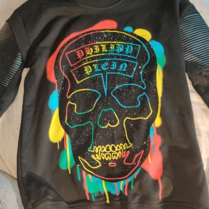 Philipp Plein svart sweatshirt med döskalle - Svart sweatshirt från Philipp Plein med stor döskalle framtill i färgglada toner och glittriga detaljer. Tröjan har ribbade muddar, coola paneler på ärmarna och tryck i rött, blått, gult och grönt. Perfekt för dig som gillar streetstyle och vill sticka ut.