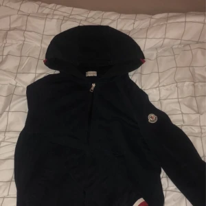 Svart hoodiejacka från Moncler M - Svart hoodiejacka från Moncler med dragkedja framtill och klassisk logga på ärmen. Jackan har huva och ribbade muddar med rödvit detalj. Perfekt för dig som vill ha en stilren och sportig look med premiumkänsla. NFC Scan på armen också, äkta vara💯