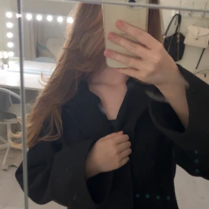 Svart kappa med bred krage - Stilren svart kappa med bred krage och knäppning framtill. Jackan har en rak passform och långa ärmar, perfekt för dig som gillar en klassisk look. Materialet känns mjukt och ger en snygg siluett. Passar dig som vill ha en enkel men trendig jacka.