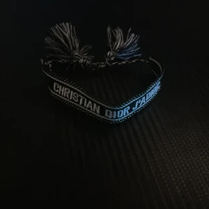 Dior Armband - 👀👀LIMITED 👀👀. Snyggt svart tygarmband från Christian Dior med texten 'CHRISTIAN DIOR J'ADIOR' broderad i vitt. Armbandet har tvinnade band och tofsar i ändarna för en cool och trendig look. Perfekt accessoar för att lyfta din outfit.