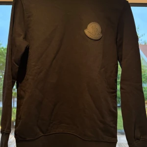 Svart sweatshirt från Moncler - Svart sweatshirt från Moncler med diskret logga på bröstet och ärmen. Tröjan har rund halsringning, ribbade muddar och är tillverkad i mjuk bomull. Perfekt för en clean och stilren look.