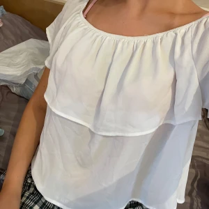 Vit volangblus från Stradivarius - Supersöt vit off shoulder blus från Stradivarius med volangdetalj över bröstet och korta ärmar. Blusen har en luftig och flowy passform med rundad halsringning och är gjord i ett tunt, lätt material som känns perfekt för varmare dagar.