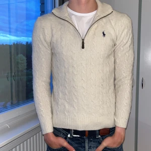 Vit ull half zip-tröja Polo Ralph Lauren - Vit kabelstickad tröja från Polo Ralph Lauren med half zip och broderad logga på bröstet. Tröjan har lång ärm, ribbade muddar och klassisk krage. Perfekt för dig som gillar en clean och stilren look. Storlek s passar även xs modell på bilden är 176 cm. Pris kan diskuteras. nypris 2800