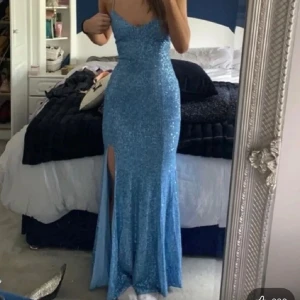 Glittrig blå långklänning med slits - Supersnygg blå långklänning med glittrigt tyg och smala axelband. Klänningen har en hög slits framtill och snörning i ryggen som ger en riktigt snygg passform. Perfekt för bal eller fest där du vill sticka ut och glänsa.🤩