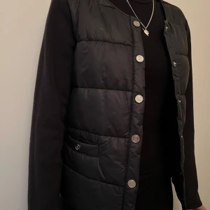 Svart dunväst från PS of Sweden - Svart dunväst från PS of Sweden med quiltad framsida, rund halsringning och silvriga knappar. Västen har två fickor framtill med knappdetaljer, däremot är en knapp på fickan borta men går ändå att använda. 