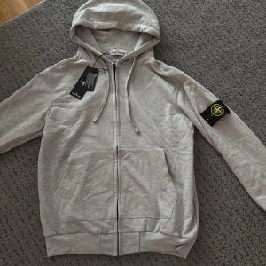 Grå hoodie från Stone Island - Snygg ljusgrå hoodie från Stone Island med dragkedja framtill, huva med snörning och klassisk logga på vänster ärm. Tröjan har känguruficka och ribbade muddar. Perfekt för dig som gillar streetwear och vill ha något stilrent men ändå ikoniskt.
