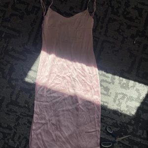 Ljusrosa långklänning med öppen rygg - Supersöt ljusrosa långklänning i satinliknande material med smala axelband och v-ringad hals. Klänningen har en snygg öppen rygg med spänne och en slimmad passform som ger en elegant siluett. Perfekt för dig som vill sticka ut med en feminin look. Den har ett litet fel på baksidan, men det är lätt att fixa. Behöver strykas.