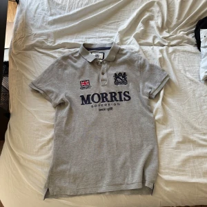 Grå pikétröja från Morris Gents - Snygg grå pikétröja från Morris Gents med broderad brittisk flagga och logga på bröstet. Klassisk krage, knappar framtill och stor Morris-text i mörkblått. Perfekt för en stilren och sportig look.