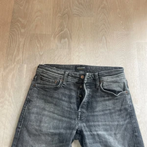 Grå jeans från Jack & Jones - Snygga grå jeans från Jack & Jones med klassisk femficksdesign och knäppning med knappar. Jeansen har en tvättad look med ljusare partier och raka ben. Perfekta för en avslappnad stil och passar till det mesta.