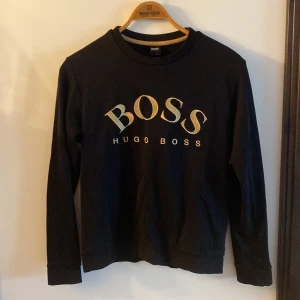 Svart tröja från Hugo Boss med logga - Svart långärmad tröja från Hugo Boss med stor guldfärgad BOSS-logga framtill. Klassisk rund halsringning och ribbade muddar. Tillverkad i mjuk bomull som känns skön mot huden. Perfekt för dig som gillar stilrena och trendiga plagg.