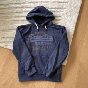 Blå hoodie från Superdry med tryck - Säljer en riktigt fet marinblå hoodie från Superdry med stort vintage-tryck i vitt och gult på bröstet. Tröjan har huva med vita snören, känguruficka och ribbade muddar. Inga defekter eller fläckar. Skriv vid minsta fråga 💯💯💯