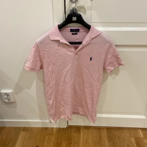 Rosa pikétröja från Polo Ralph Lauren - Snygg rosa pikétröja från Polo Ralph Lauren med klassisk krage och korta ärmar. Tröjan har den ikoniska blå loggan broderad på bröstet och är gjord i mjuk bomull. Perfekt för en avslappnad men stilren look.