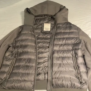 Grå pufferjacka med huva från Replay - Snygg grå pufferjacka från Replay med huva och dragkedja. Jackan har quiltad framsida och rygg i glansigt material, medan ärmarna är i mjukare tyg. Fickor med dragkedja och en extra ficka på ärmen ger en cool touch. Pris kan diskuteras 😁