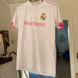 Fotbolls tröja - Vit fotbollströja från Real Madrid i storlek XS. Tröjan har rosa detaljer på ärmslut och axlar samt klubbens emblem och texten 'Real Madrid' på bröstet. Tillverkad i ett lätt och andningsbart material, perfekt för träning eller match.