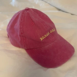 Vintagelooking keps  - Snygg rosa keps med broderad gul text framtill. Klassisk böjd skärm och justerbart spänne bak i metall. Perfekt accessoar för soliga dagar och strandhäng. Tillverkad i mjuk bomull för en bekväm känsla.