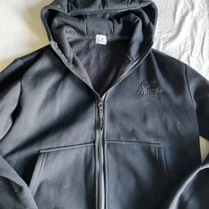 Svart hoodiejacka från C.P. Company - Svart hoodiejacka från C.P. Company med broderad logga på bröstet och dragkedja framtill. Jackan har huva med inbyggda glasögon och två stora fickor. Mjuk insida och stilren look, perfekt för dig som gillar streetwear.