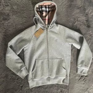 Grå hoodie från Burberry med dragkedja - Snygg grå hoodie från Burberry med klassiskt rutigt foder i huvan och broderad logga på bröstet. Tröjan har dragkedja, snörning i huvan och två stora fickor framtill. Perfekt för dig som gillar stilrena och exklusiva plagg. Passar någon som e runt 165