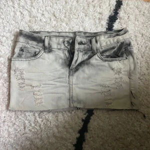 Grå jeanskjol med slitningar, stl M - Säljer en grå, kort jeanskjol i storlek M med slitna detaljer framtill och baktill. Kjolen har fem fickor, bälteshällor och rå kant nertill för en edgy vibe. Perfekt för dig som gillar streetstyle och vill ha en cool look. Skriv för fler frågor eller bilder🥰