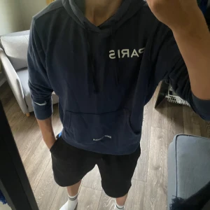 Blå hoodie Paris Saint Germain - Snygg marinblå hoodie från Nike x Paris Saint Germain med vit text på bröstet. Klassisk känguruficka, huva med snörning och långa ärmar. Perfekt för dig som gillar sportig och avslappnad stil.