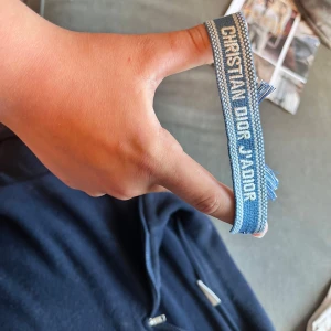 Blått Christian Dior J'Adior armband - Snyggt blått armband från Christian Dior med texten 'J'Adior' invävd i vitt. Armbandet är gjort i tyg med justerbar knytning och fransar i ändarna. Perfekt accessoar för att lyfta din outfit med en exklusiv touch.