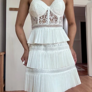 Vit spetsklänning från ASOS Design - Superfin vit kortklänning från ASOS Design med spetsliv, plisserade volanger och smala axelband. Klänningen har v-ringad byst, genombruten spets och dragkedja i sidan. Perfekt för dig som vill sticka ut med en romantisk och trendig look.