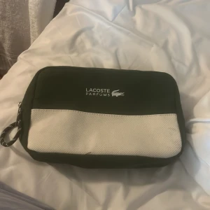 Grön och vit necessär från Lacoste - Snygg necessär från Lacoste Parfums i mörkgrönt och vitt tyg. Praktisk rektangulär form med dragkedja och coolt Lacoste-berlock på zipen. Perfekt storlek för att ha med sig essentials och enkel att bära med handtag i sidan.