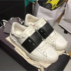 Valentino Open - Snygga Valentino Open sneakers i vitt skinn med bred svart läderdetalj över vristen. Klassisk rund tå, vita snören och coola nitar på hälen. Perfekt för dig som gillar stilrena och lyxiga sneakers med en twist. Storlek 