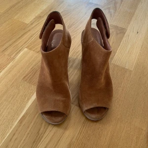 Bruna mockasandaler med klack - Snygga sandaler i brun mocka med öppen tå och bred klack. Skorna har en stilren design med rundad form och bred rem runt hälen. Perfekta för dig som vill ha en trendig och bekväm sko med lite höjd. Använda en enda gång 