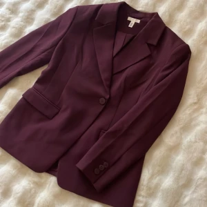 Vinröd kavaj från H&M - Vinröd kavaj från H&M med klassisk krage, två fickor med lock och enkelknäppning framtill. Snygg, stilren modell som passar perfekt till kostymbyxor eller jeans. Materialet är slätt och känns bekvämt mot huden.