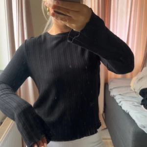 Svart ribbad långärmad tröja - Svart långärmad tröja med ribbad struktur och lätt utsvängda ärmslut. Tröjan har en enkel rund halsringning och är i ett mjukt, stretchigt material som sitter snyggt på kroppen. Perfekt till jeans eller kjol för en clean look.