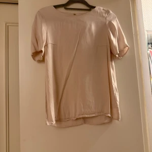 Beige blus i sidenlook med kort ärm - En stilren beige blus med rund halsringning och korta ärmar. Blusen har en lätt glansig yta som påminner om siden och en snygg öppning i ryggen. Perfekt för dig som gillar en enkel men elegant vibe.