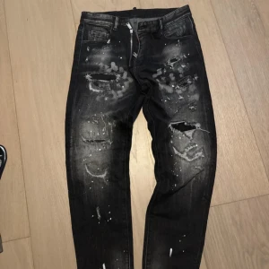 Svarta distressed jeans från Dsquared2 - Snygga svarta jeans från Dsquared2 med coola slitningar, hål och vit färgstänk över hela byxorna. Modellen har klassisk femficksdesign och smal passform. Perfekta för dig som gillar en edgy streetstil.