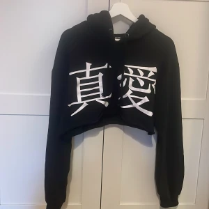 Svart croppad hoodie från Bik Bok - Svart croppad hoodie från Bik Bok med stora vita japanska tecken framtill och texten 'TRUE LOVE' på huvan. Tröjan har huva med snörning och långa ärmar. Perfekt för en chill och trendig look. Ärmlängd 57 cm, 29 cm i längden. 